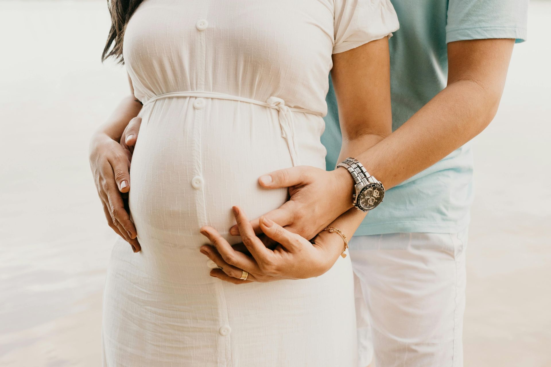Crop anonymous young man in casual clothes embracing belly of pregnant wife while standing on seashore