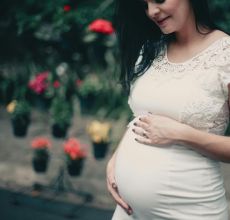Pregnant woman in white lace dress, posing outdoors with flowers, exuding happiness.