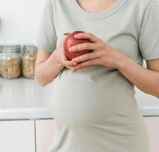 Unrecognizable pregnant female with ripe red apple for healthy diet in hands standing near counter in light kitchen at home