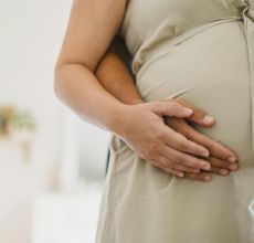 Crop faceless male holding hands on pregnant female tummy while standing together in light room