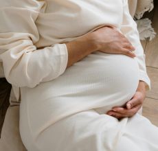A serene moment of a pregnant woman in a cozy white dress holding her belly.