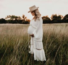 Stylish pregnant woman in a white dress and hat standing in a grassy field during sunset.